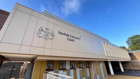 Norfolk Coroner's Court building, clad in a gold finish, with black lettering identifying the building with His Majesty's crown emblem featured to its left. the lower part of the building features mustard yellow and white panels with dark framed windows. The sky above is blue with wisps of white cloud.