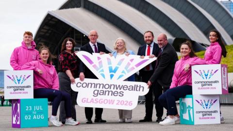 Athletes in pink hoodies flank official figures who are standing outside the SEC Armadillo in Glasgow holding Commonwealth Games branding. They are, from left, Glasgow City Council leader Susan Aitken; First Minister John Swinney; Commonwealth Games Federation chief executive Katie Sadleir; Scottish Secretary Ian Murray and the chief executive of Commonwealth Games Scotland Jon Doig.