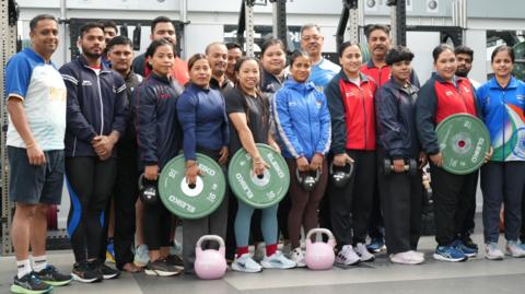 The men and women of the Indian weightlifting team lined up in a gym to pose for a photo, They are wearing gym clothes and some are holding large, round weights.