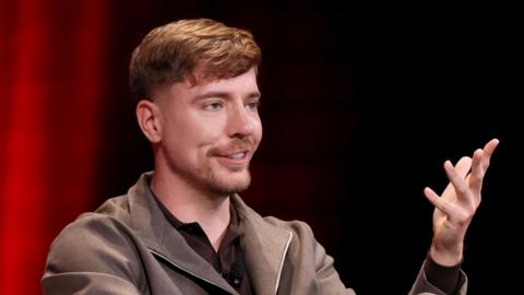 MrBeast, real name Jimmy Donaldson, at an event. He is wearing a brown t-shirt and zip top, with his left arm gesturing as he speaks.