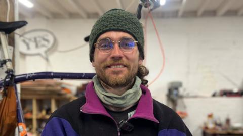Alan is smiling at the camera. He is standing in a workshop with his tall bike behind him. He is wearing round glasses, a green beanie hat and a colourful jumper.