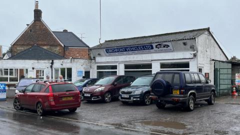 Forecourt of Urchfont Vehicles garage with four cars near a road.