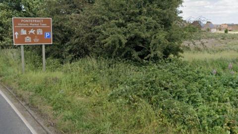 Fields beside a road with a brown sign stating Pontefract, Historic Market Town
