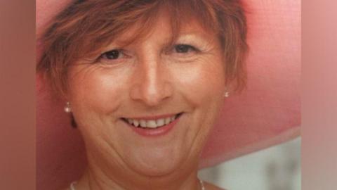Christine Jones, with short red hair and wearing a wide brimmed pink hat, smiles at the camera.