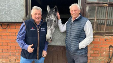 Paul Nicholls (left) standing with his arms by his side and his thumb up in his right hand, Chris Barber (right) standing with one hand in his pocket and with his right arm up touching the head of horse (centre) No Drama This End whose head is coming out of the stable