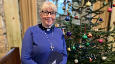 Reverend Lisa Coupland stood in front a decorated Christmas tree. She is holding a blue book in both hands and is wearing a blue top and black and white collar. She's also wearing a silver necklace, black glasses and is smiling at the camera. Behind her is a brick wall and a window at the centre with two long curtains either side of it. 