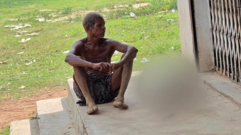 A man wearing a dark printed dhoti - which is a cloth tied around the waist and covering until the knee - sits on stairs. With him are the remains of his sister - which have been blurred. There is green grass in the background and a metal gate on one side. 