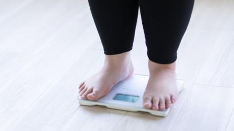 A child stands on weighing scales wearing black leggings and bare feet