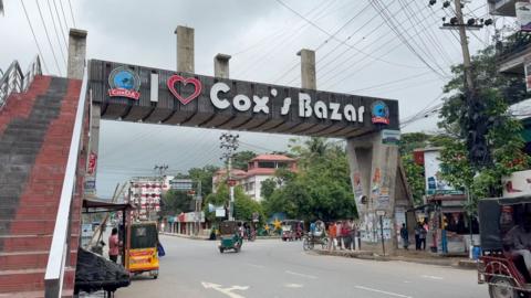 A street view featuring a large overhead sign that reads “I ♥ Cox’s Bazar” in bold letters. The sign is supported by concrete pillars and has logos for local development authorities. Below, the road has light traffic with auto-rickshaws and pedestrians. Surrounding buildings display colorful signage, and electrical wires are visible overhead against a cloudy sky.