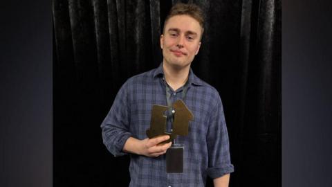 Image of a Sam Fender, from the waist up. He has short dark hair, stubble and a half smile and is wearing a blue checked shirt. In his right hand he is holding an award, two metal shapes on a black base.