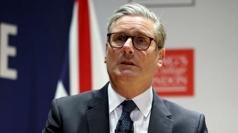 Keir Starmer looks on as he speaks at an event in London on Thursday. He's wearing a suit and tie with glasses on.