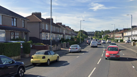 Residential street with terraced houses, parked cars and moving traffic under a clear blue sky