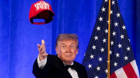 Trump in bow tie, before American flag, tosses red "Trump 2028" cap in air