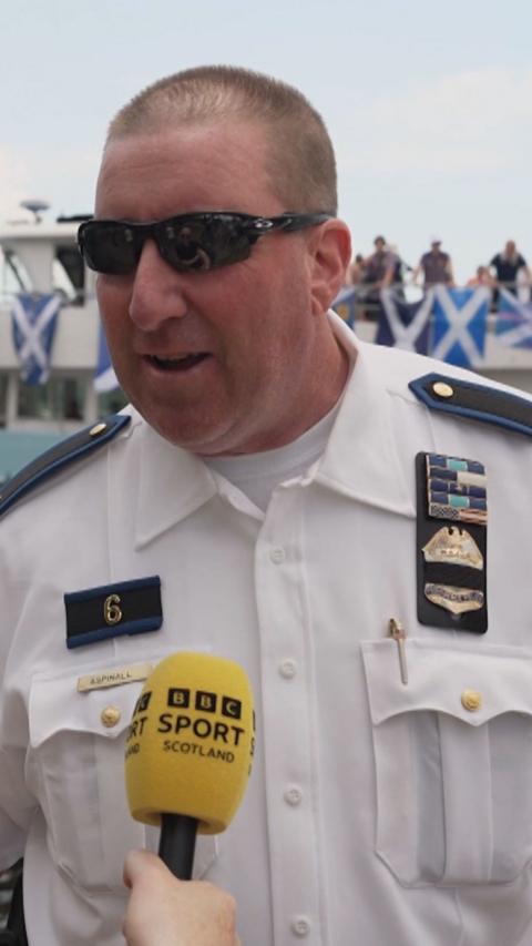 A police officer in his uniform and black sunglasses is speaking into a yellow 'BBC Sport Scotland' branded microphone.