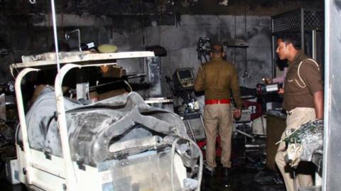 Two male police officers, one with a torch, examine burned medical equipment in a fire-blackened room at Maharani Laxmibai Medical College.