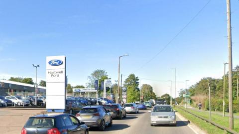 Redbourn Road, Hemel Hempstead, with cars on the road, by a car dealership. There are trees and grass to the left, and signs for the dealership to the right. 