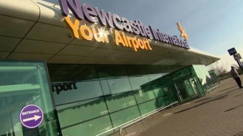 The outside of Newcastle Airport which has a glass frontage and an orange and purple sign saying Newcastle International - Your airport