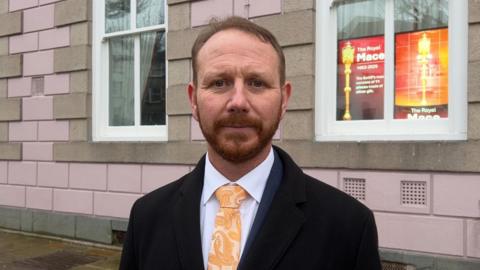 Tom Coles is stood in front of the States Assembly building. He is wearing a black jacket and yellow tie