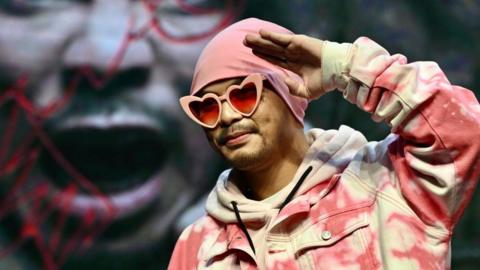 Namewee wearing a pink beanie, pink heart-shaped sunglasses and a pink denim jacket, raising his hand to his forehead in a saluting pose.