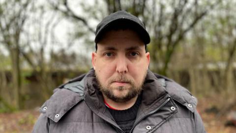 James Fenton looking at the camera. He has a beard and is wearing a padded a black padded coat and a black baseball cap.