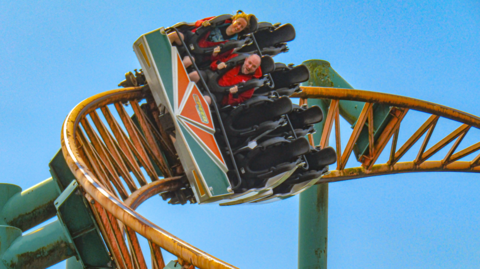 An orange and blue rollercoaster track, with sky behind. There is a rollercoaster cart on the track, with a woman and a man, both wearing red, strapped into the six-seater cart. They are grasping their hands onto the safety vests and smiling. 