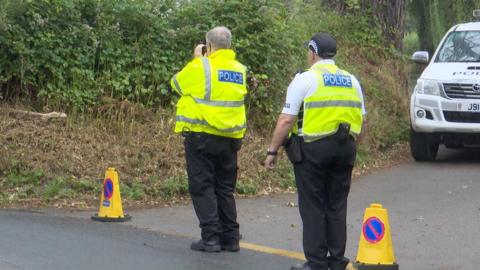 Two people in high vis jackets marked with the word Police stand with their  back to the camera. One of them appears to be using binoculars to look into a hedge in front of them. They are in a lay by and there are two yellow traffic cones near them. A four by four marked Police is parked to the right.