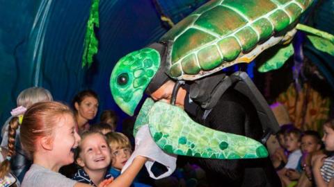 A performer wearing a sea turtle puppet interacting with a group of children inside a dimly lit inflatable whale's stomach. The children, who are sitting on the floor, look happy. One girl is reaching out to touch the puppet’s fin. 
