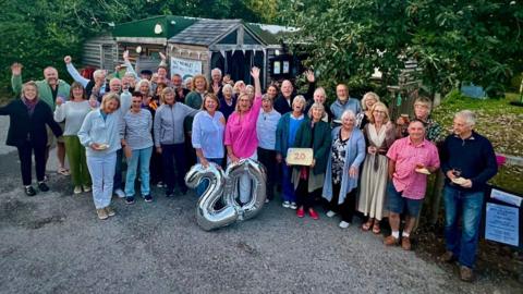 A group of volunteers are gathered out side the community ship cabin with signs that say 20 celebrating its 20th birthday. They are all smiling and looking at the camera with the cabin behind them amongst threes and bushes.
