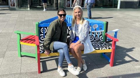 Two young women sit on a colourful bench