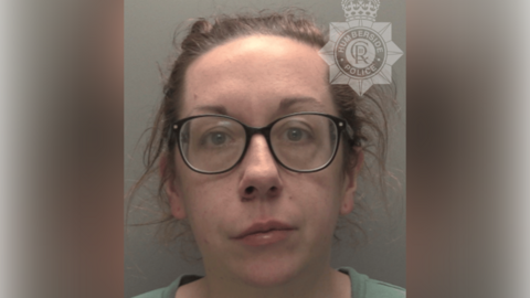 A police custody shot of Gemma Phoenix looking at the camera. She has brown hair tied back and is wearing a green t-shirt and black-rimmed glasses. The wall behind her is grey.