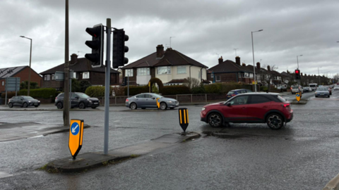 A number of cars are passing through a junction with traffic lights. Semi-detached houses can be seen.