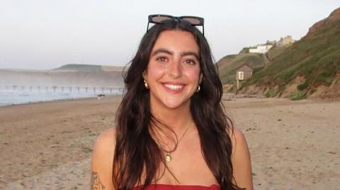 A young woman standing on a sandy beach, facing the camera wearing a strapless red dress. She has long dark hair and is wearing sunglasses on top of her head.