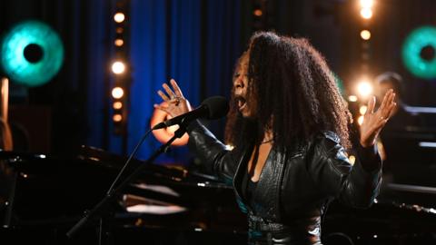 Beverley Knight, singing with her hands spread wide, wearing a leather jacket and black t-shirt