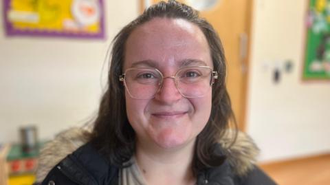 A smiling Anna looks directly at the camera as she is photographed inside a family hub. She is wearing glasses and has dark hair, and is wearing a dark jacket with a fur-lined hood. Behind her you can see brightly coloured wall art. 