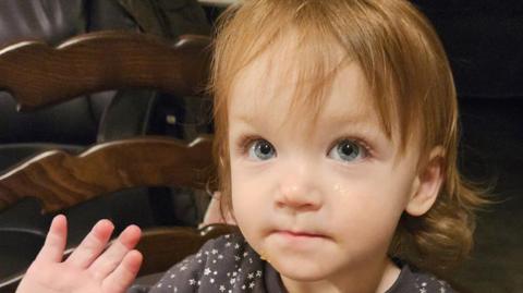 A two-year-old girl with shoulder-length brown hair and blue eyes is wearing a dark top with white stars on it. She is sitting on a chair and is holding up her right hand