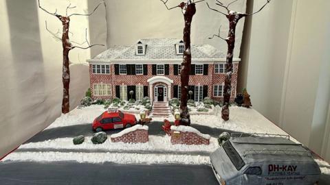 The cake in the window it shows the two-storey red brick Home Alone house with icing snow on the roof and front lawns on the driveway is a confectionary car and van in the road