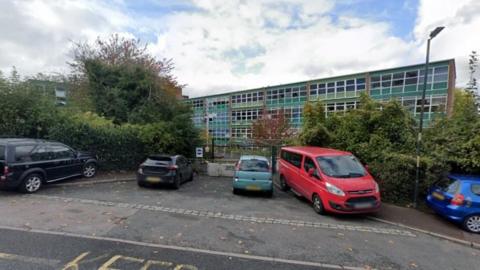 A green school building covered in windows stands behind a car park filled with vehicles
