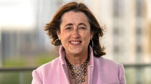 Jane Dodds who has brown hair and a necklace made out of hexagons in a pink coat