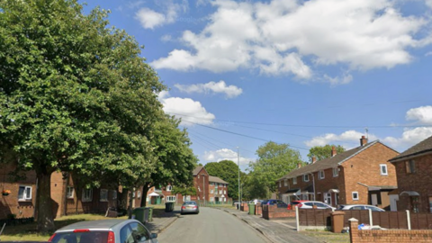 Large trees can be seen on the left of the street which has houses either side with cars parked in the road or on some drives. The sky is blue with some clouds.