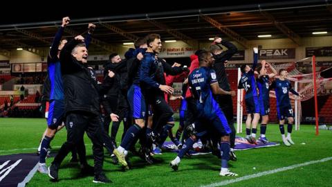 Lincoln City players and coaching staff wearing dark blue kits gather on the pitch near a goal, with several people jumping and raising clenched fists. Empty stadium seats, advertising boards and floodlit stands are in the background.