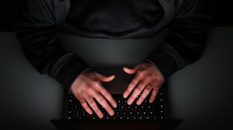 This darkened images shows a view from above of a persons hands working on the keyboard of a computer. The person is in shadow and they are wearing a black jumper. 