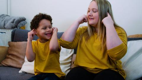 Mother and son playing together and smiling, both are pointing to their ears.