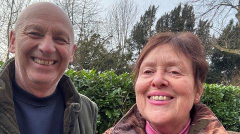 A man and a woman are smiling at the camera with a hedge behind them. Both are wearing coats. He has a blue jumper and she has a pink jumper.