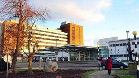 The front entrance of Altnagelvin hospital in Londonderry. A woman in a red coat can be seen walking past a statue while a number of parked cars sit outside the hospital entrance. It is dry and sunny, two trees are in the foreground, a number of hospital buildings in the background.