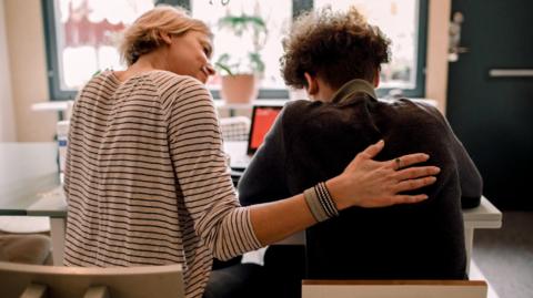 Stock photo of mother, wearing a stripy top, with short blonde hair, sitting by teenage son, with brown curly hair, studying at home - at a table, in front of a laptop.