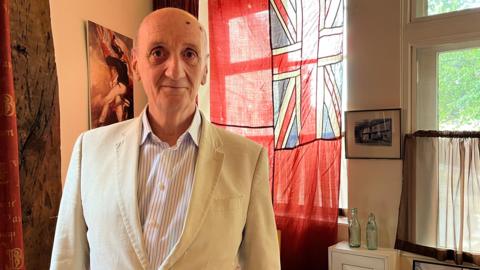 Peter Berthoud stands in front of a red flag with a union flag in its top corner. The room he is in has old wooden beams holding up the roof. Peter is bald, wearing a cream linen suit and white striped shirt. 