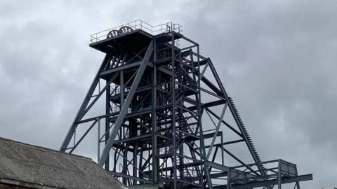 South Crofty tin mine. A grey metal structure is suspended above the ground with ladders/steps leading to the very top. The sky is also grey.