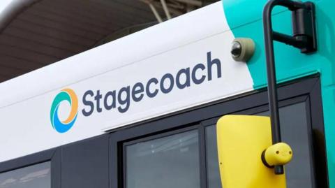 The right side of a white and mint green bus with a yellow wing mirror. The image is zoomed in above the driver's window showing the top of the window and the word Stagecoach written in black letters on the bus itself