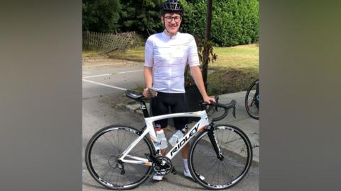 Jack stood behind his white road bike. Jack is wearing black shorts, a white cyclist top, clip-in shoes, and a helmet. He is wearing glasses and is smiling to camera.