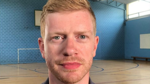 A man with ginger hair and a ginger beard in a sports hall with a wooden floor and blue walls
